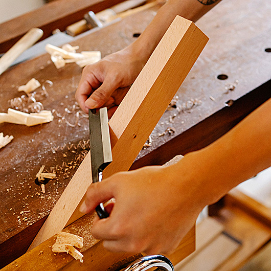 Hände mit Ziehmesser bearbeiten ein Stück Holz
