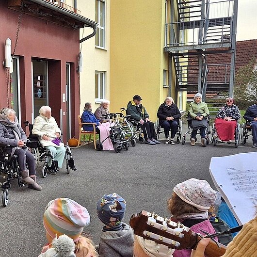 Senioren sitzen im Halbkreis vor einer Gruppe singender Kinder