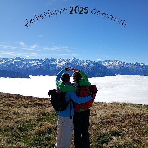 Zwei Personen vor verschneiten Bergen halten sich im Arm und bilden mit den Händen ein Herz. 