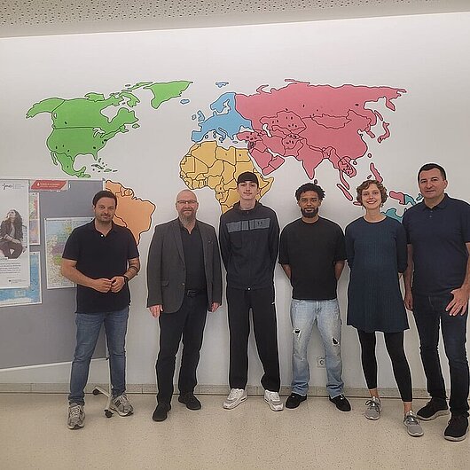 Es stehen 6 Personen vor einem Weltkarten Bild an der Wand